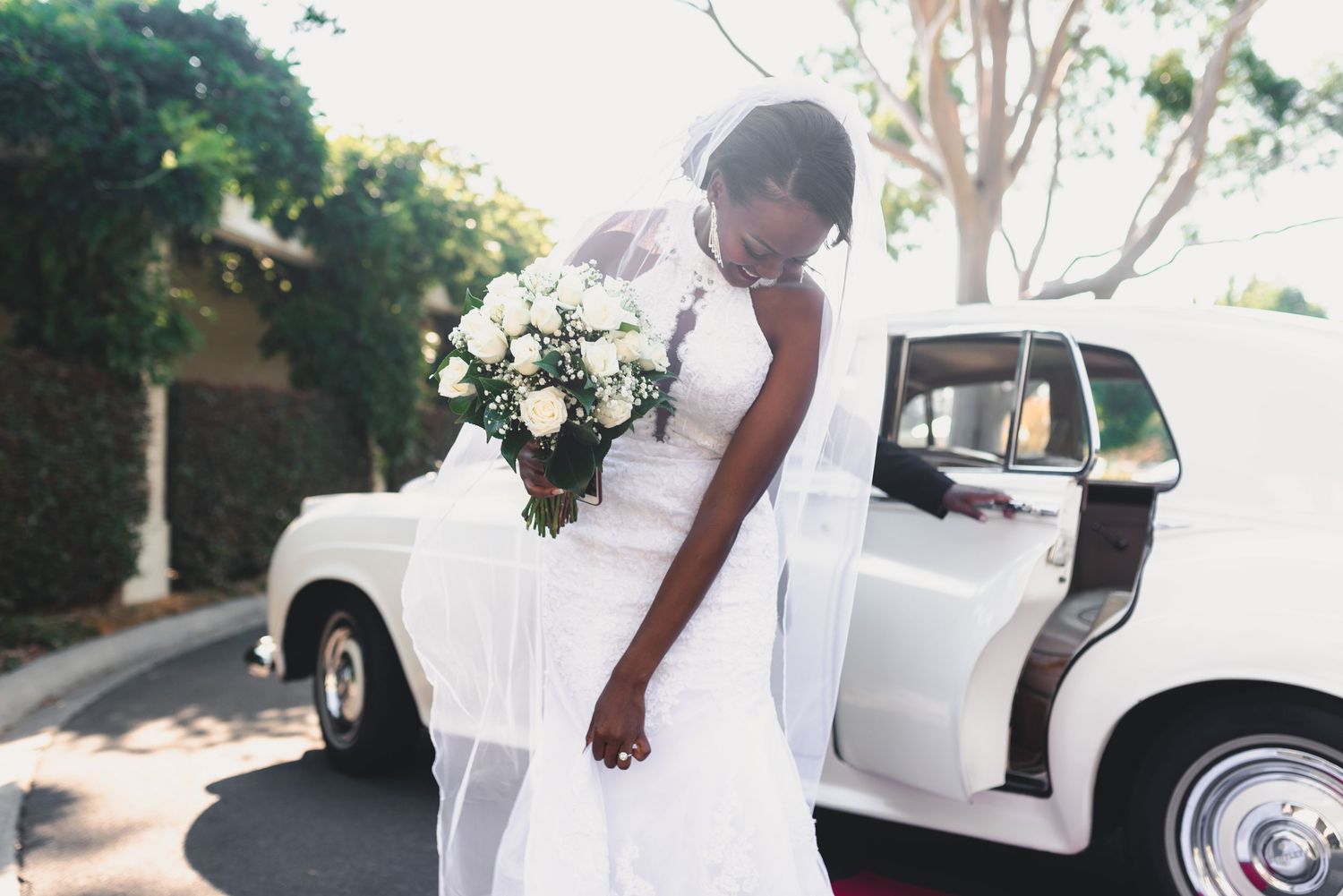 Elegant bride in white dress holding bouquet next to vintage white car.