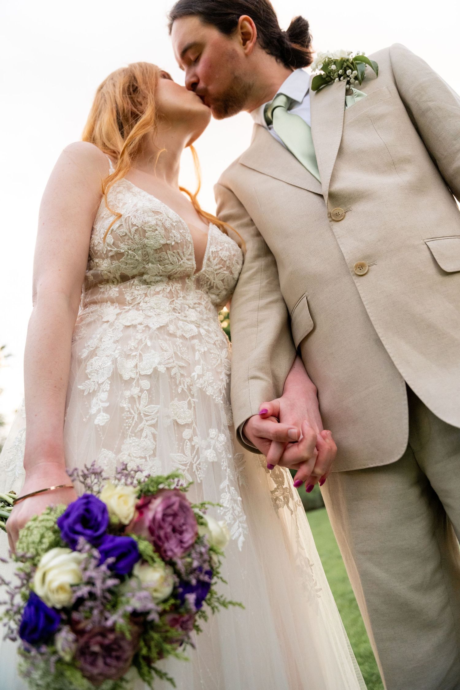 Close-up details of a bride in lace dress and groom in beige suit holding a purple and white bouquet.