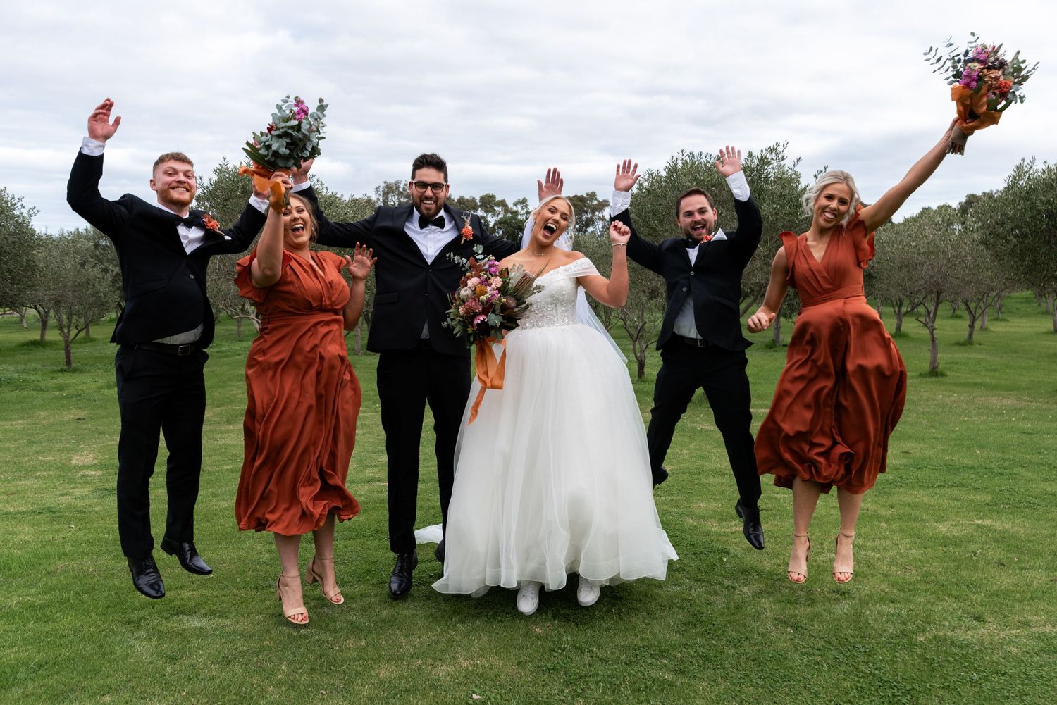 Wedding party in black suits and rust-colored dresses jumping joyfully on a green lawn under cloudy skies.