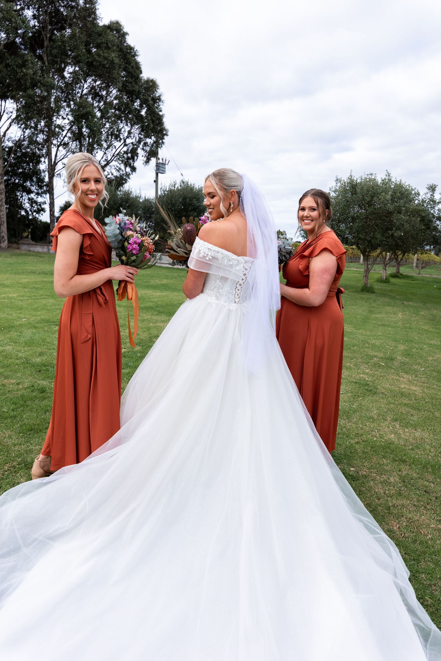 Bridesmaids in rust-colored dresses help arrange a flowing white wedding gown on a grassy lawn.