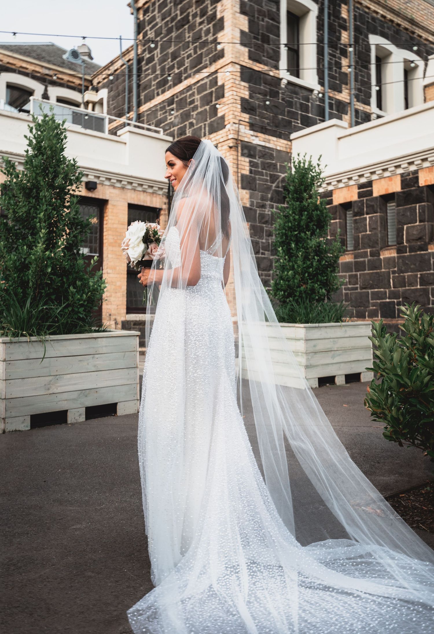 Stunning cathedral-length veil with delicate lace details trails behind an elegant white wedding gown on stone courtyard.