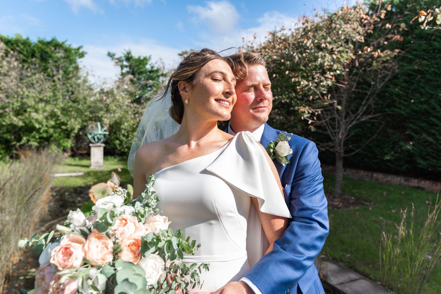 Newlyweds share intimate moment in garden setting with peach and white floral bouquet.