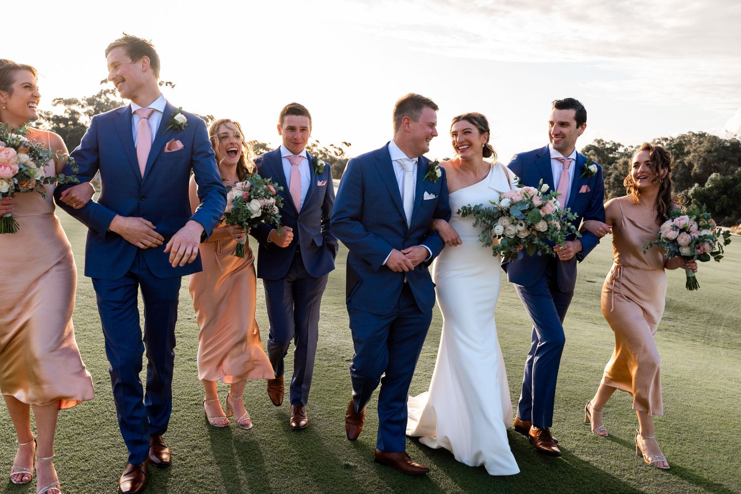 Wedding party in navy suits and blush dresses walking together on grass during outdoor celebration.