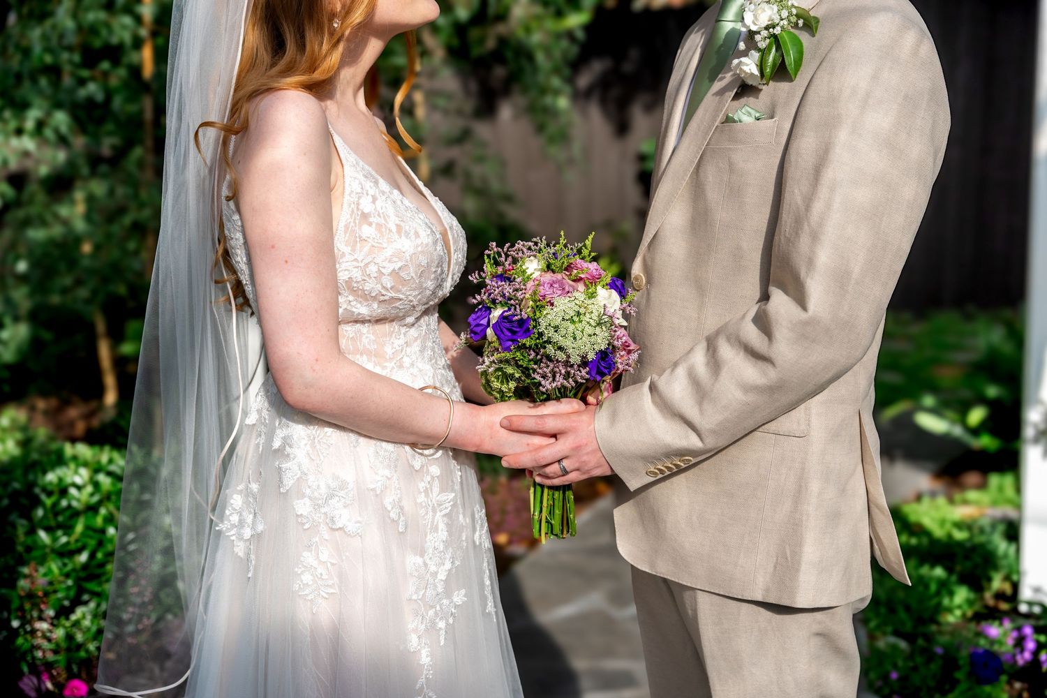 A couple holds a vibrant purple and white wedding bouquet together against a garden backdrop.
