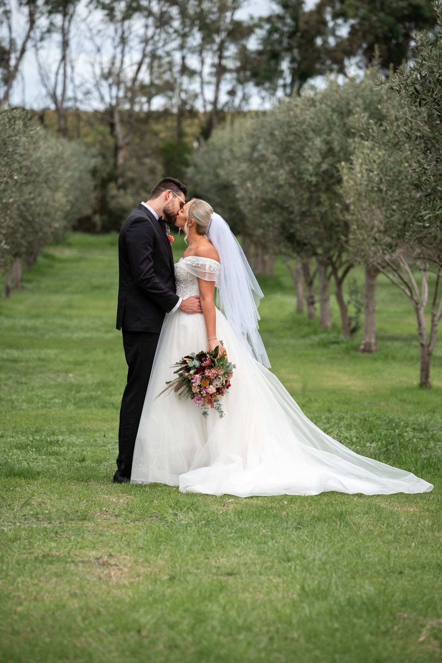 A romantic wedding couple shares an intimate moment in an olive grove garden with a flowing white dress and bouquet.