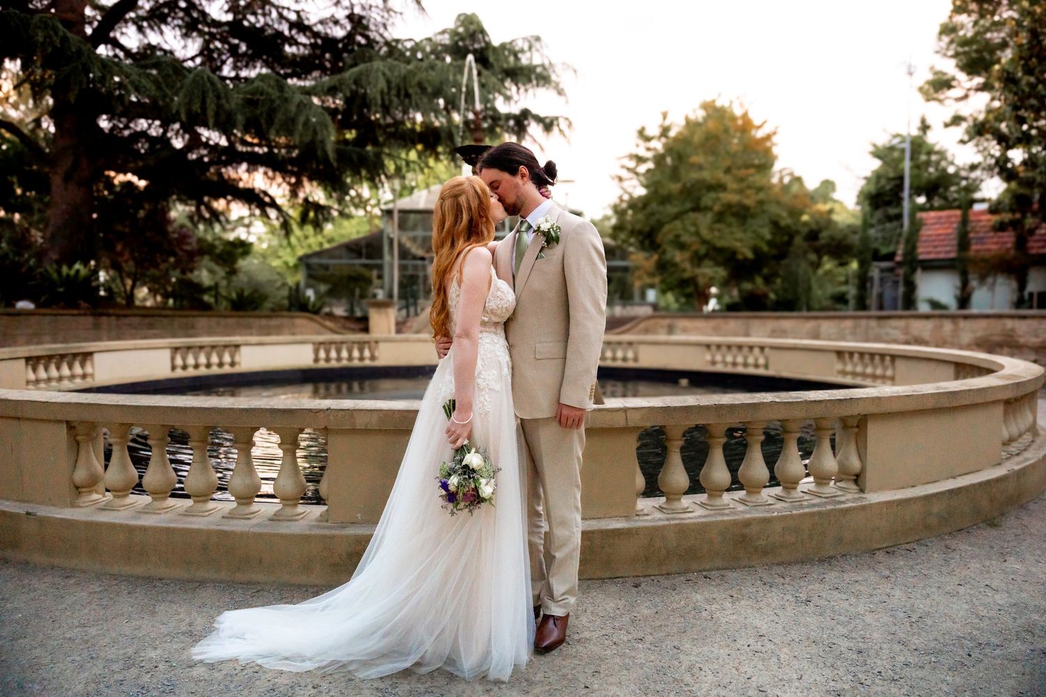 A romantic wedding couple stands on a stone balcony overlooking a garden during a sunset ceremony.