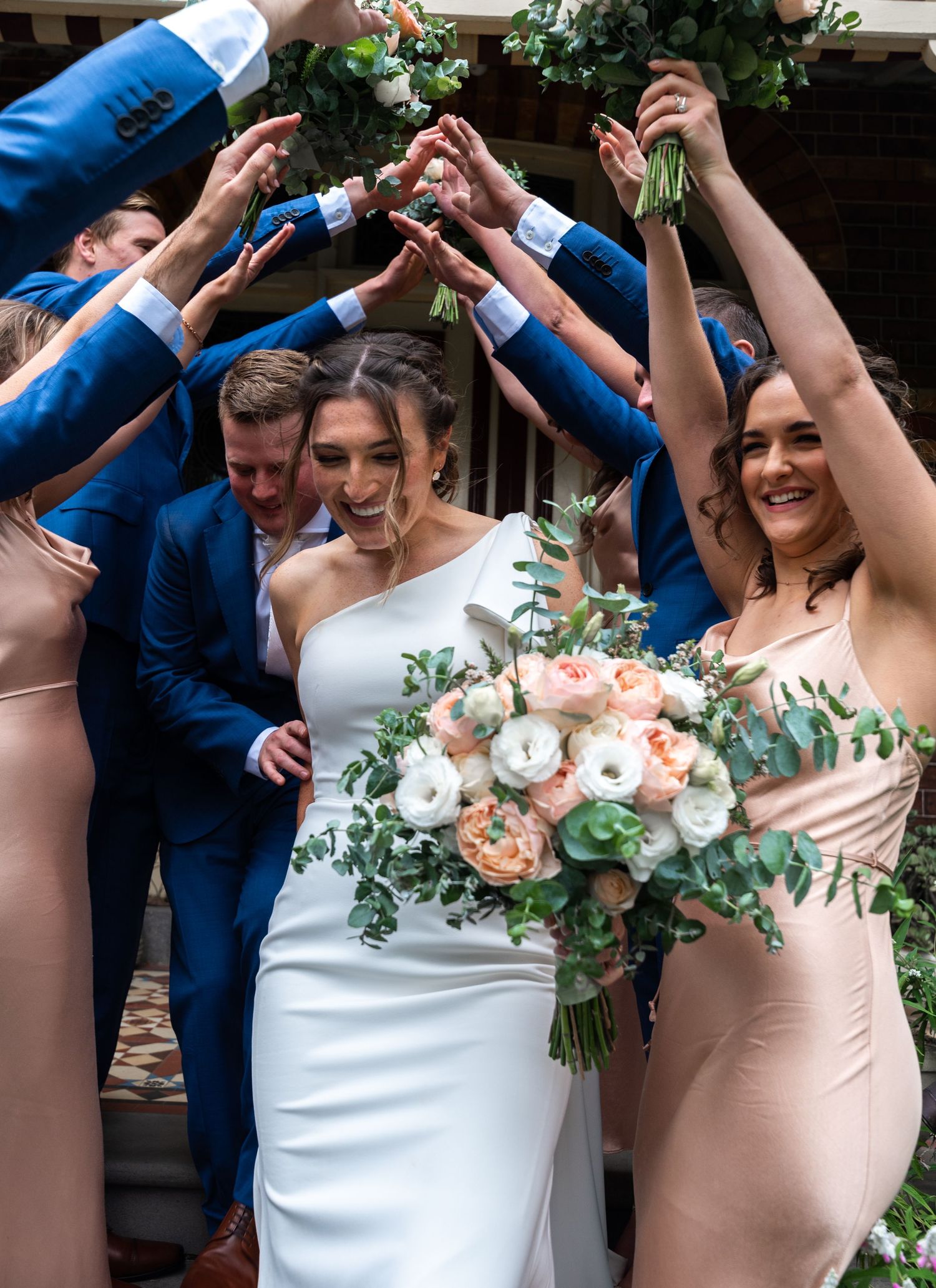 Wedding party in blue suits and pink dresses joining hands in celebration outdoors.