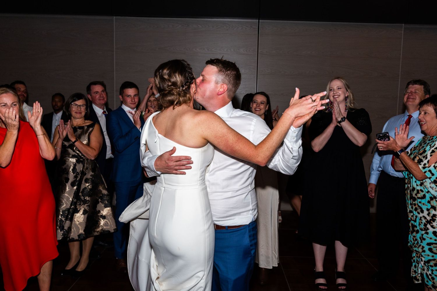 Wedding couple shares their first dance surrounded by guests in a dimly lit reception hall.