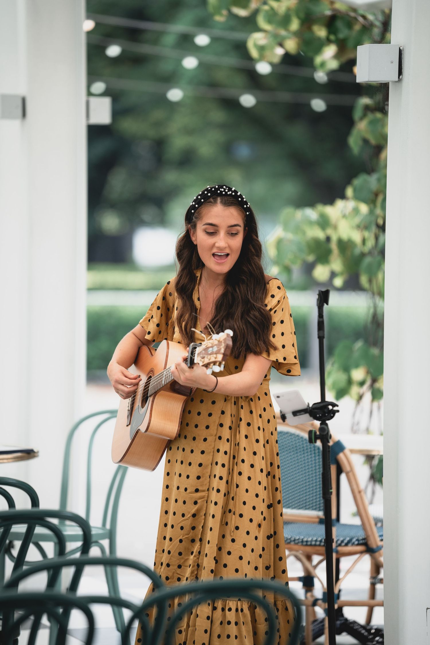Musician in polka dot dress performs with acoustic guitar on outdoor patio with string lights and chairs.