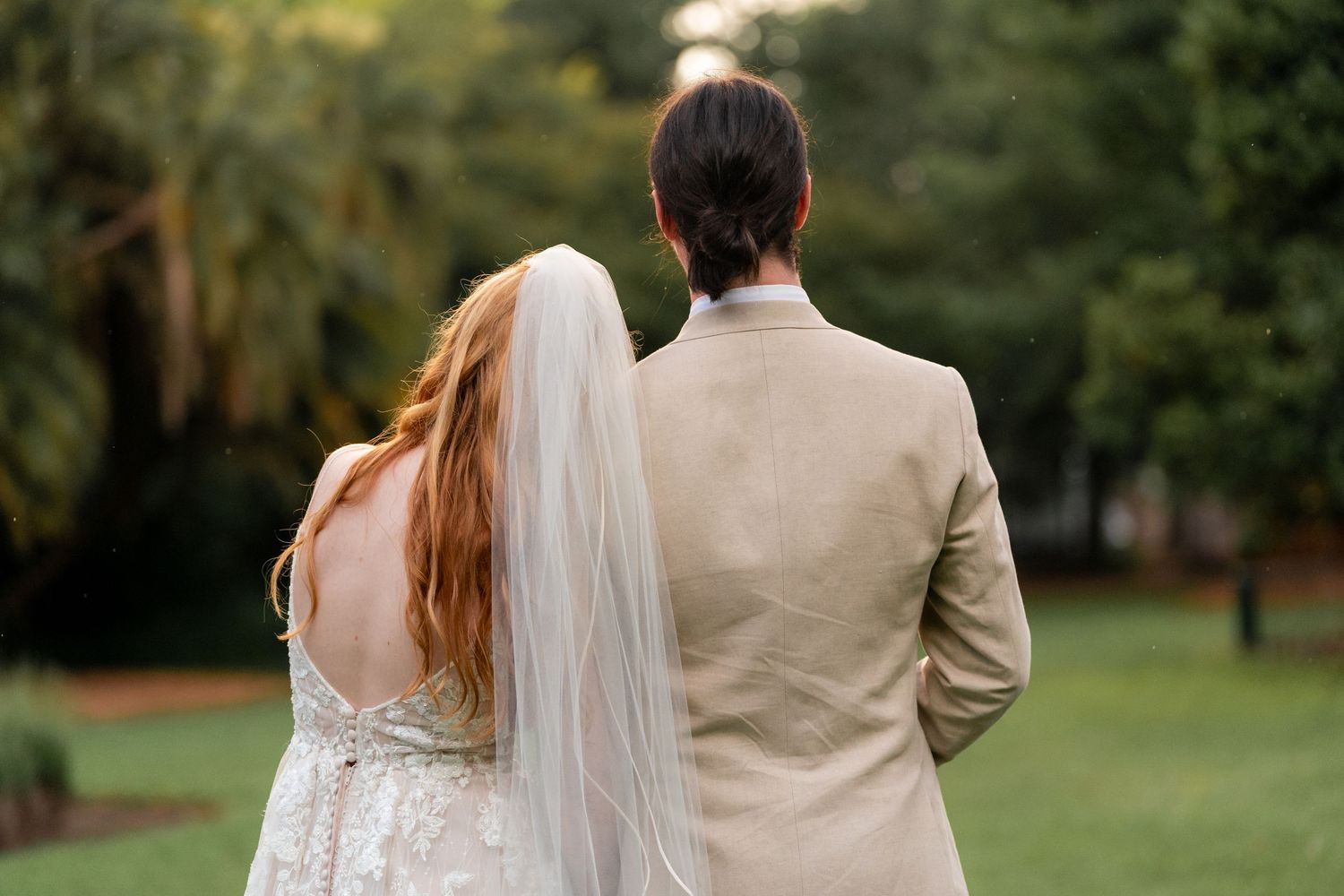 Back view of newlyweds standing together outdoors, bride in white lace dress with veil and groom in beige suit.
