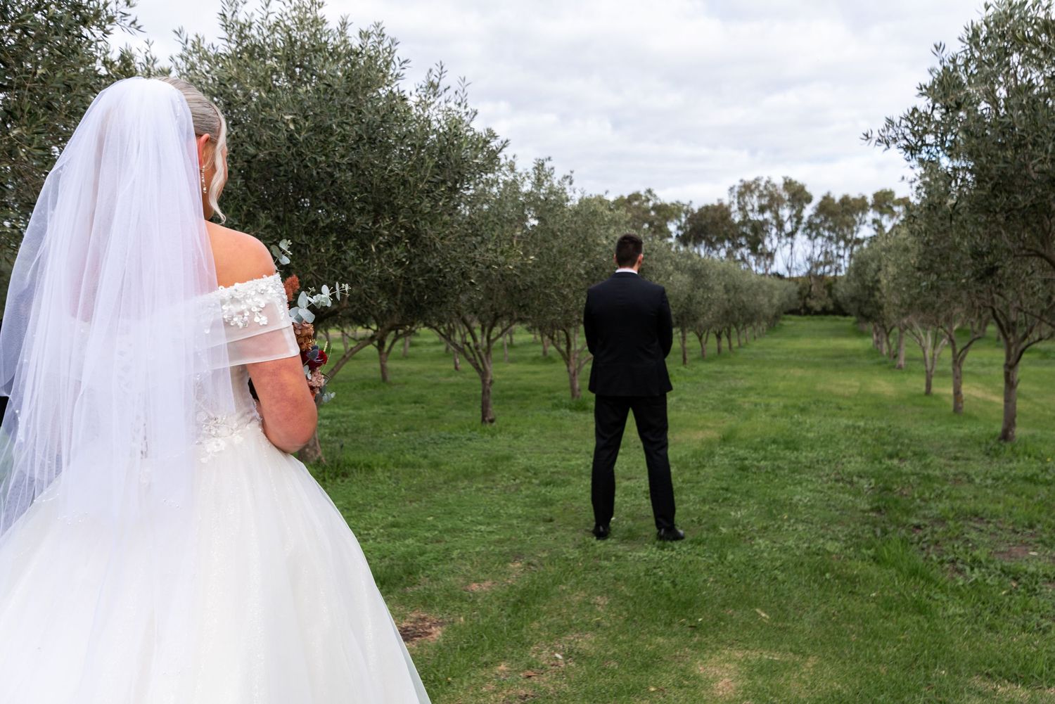 A bride in a white wedding dress walks through a green orchard towards someone in a black suit in the distance.