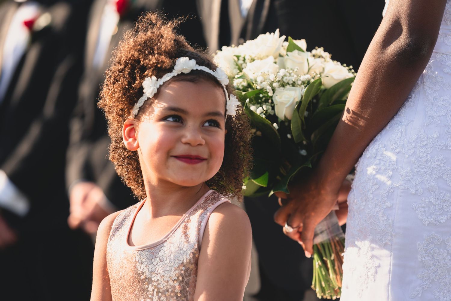 A young flower girl in a white dress smiles while holding a bouquet at a wedding ceremony.