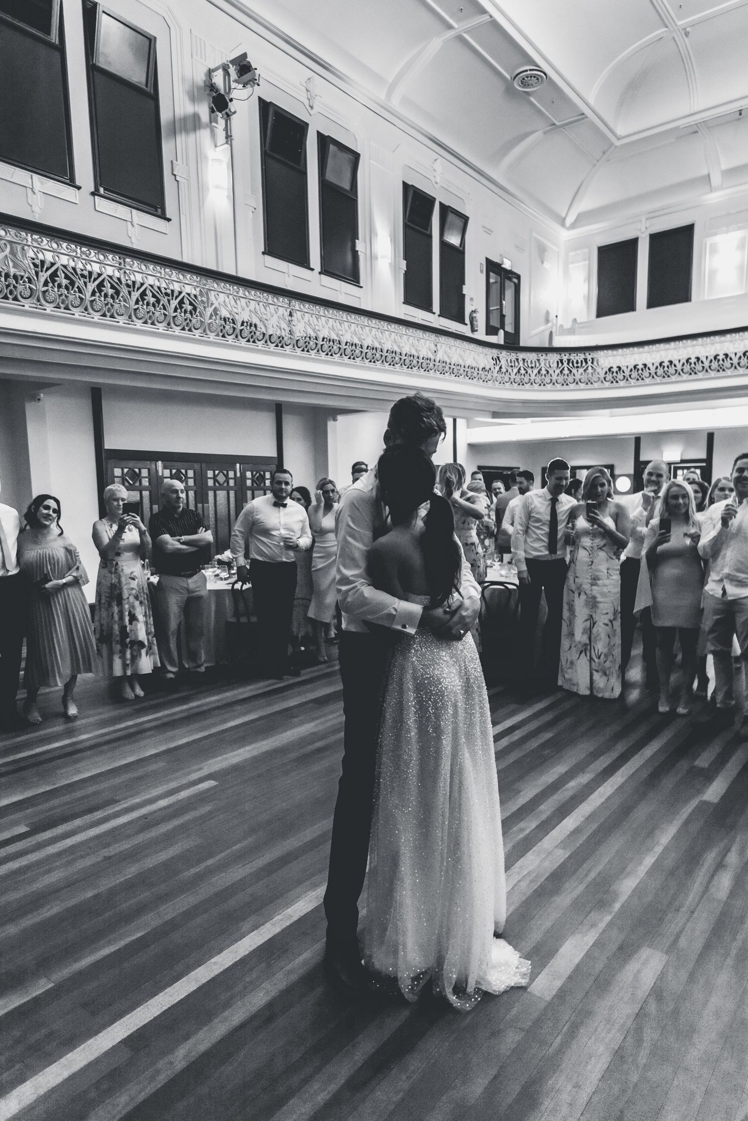 A black and white photograph captures a couple's first dance at their wedding reception in a vintage ballroom with hardwood floors.
