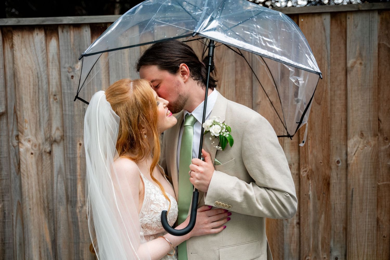 A romantic couple shares an intimate moment under a clear umbrella in front of a rustic wooden wall.
