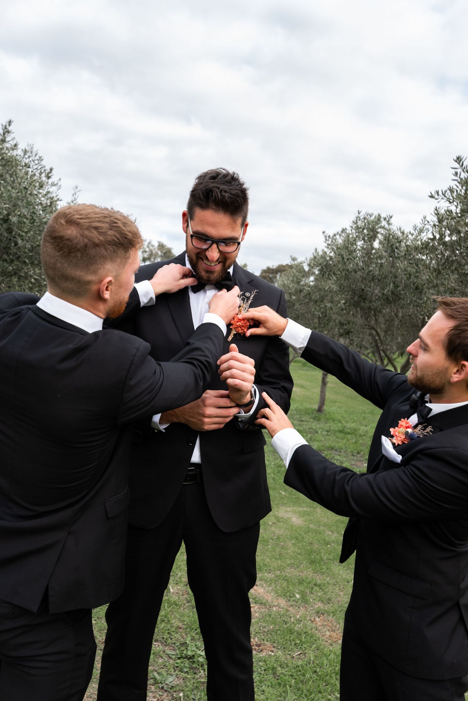 Groomsmen in black suits playfully help each other adjust their ties and boutonnieres in a candid photo sequence outdoors.