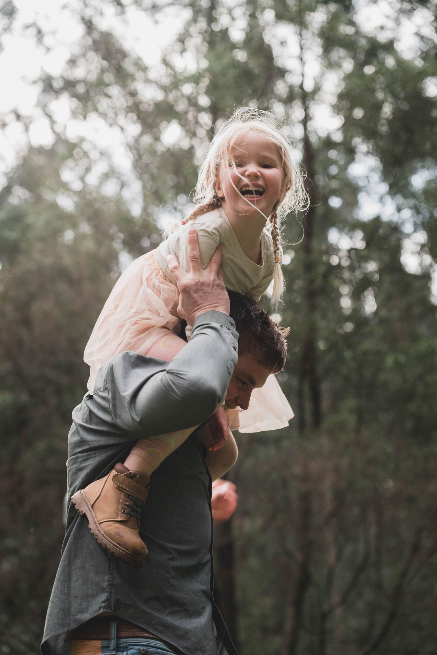 A couple embraces joyfully in a wooded outdoor setting, with the person wearing a grey shirt lifting their partner.