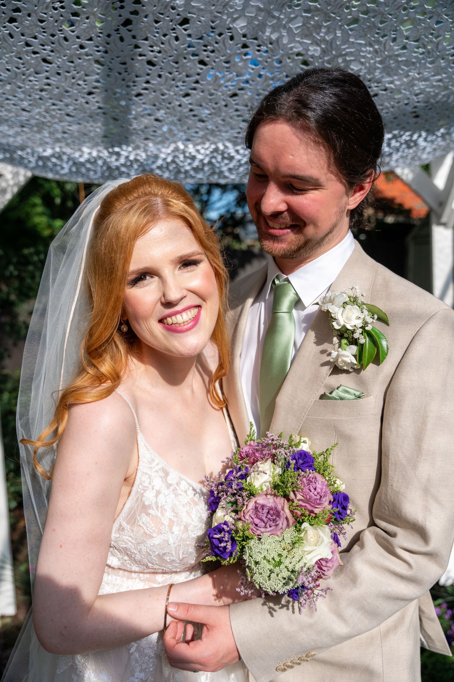 A joyful wedding couple shares intimate moments while holding a purple and white floral bouquet.