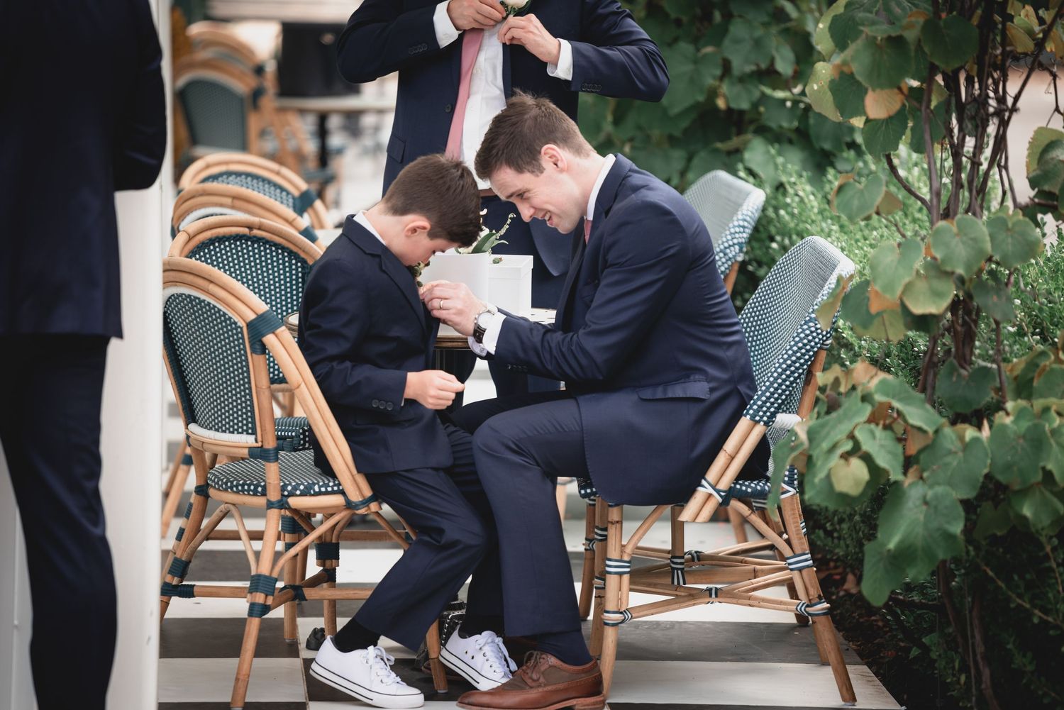 Person in a navy suit sits at an outdoor cafe table while looking down at something in their hands.