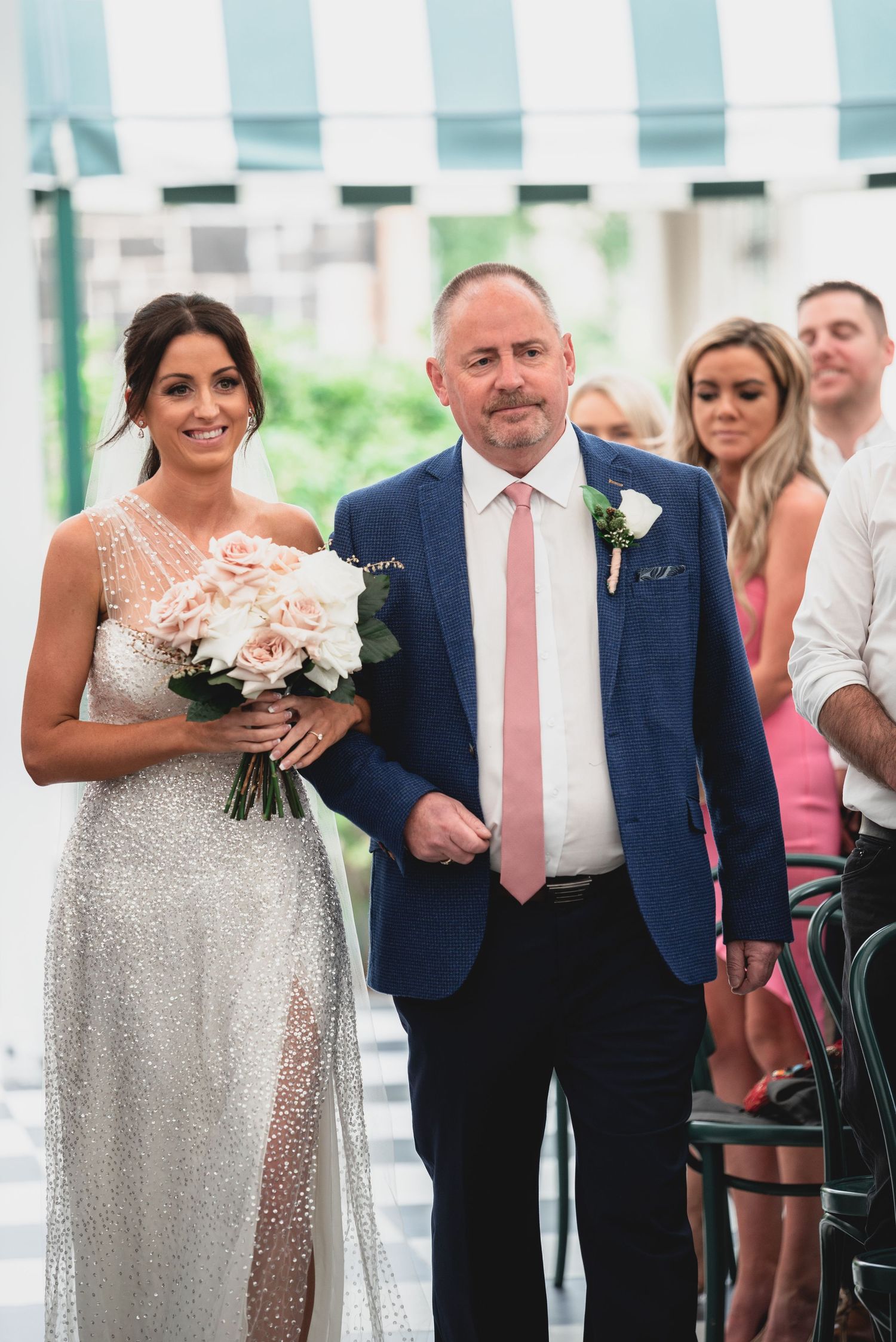 Wedding party members in navy suits and pink ties stand alongside bridesmaids in sparkly silver dresses holding bouquets.