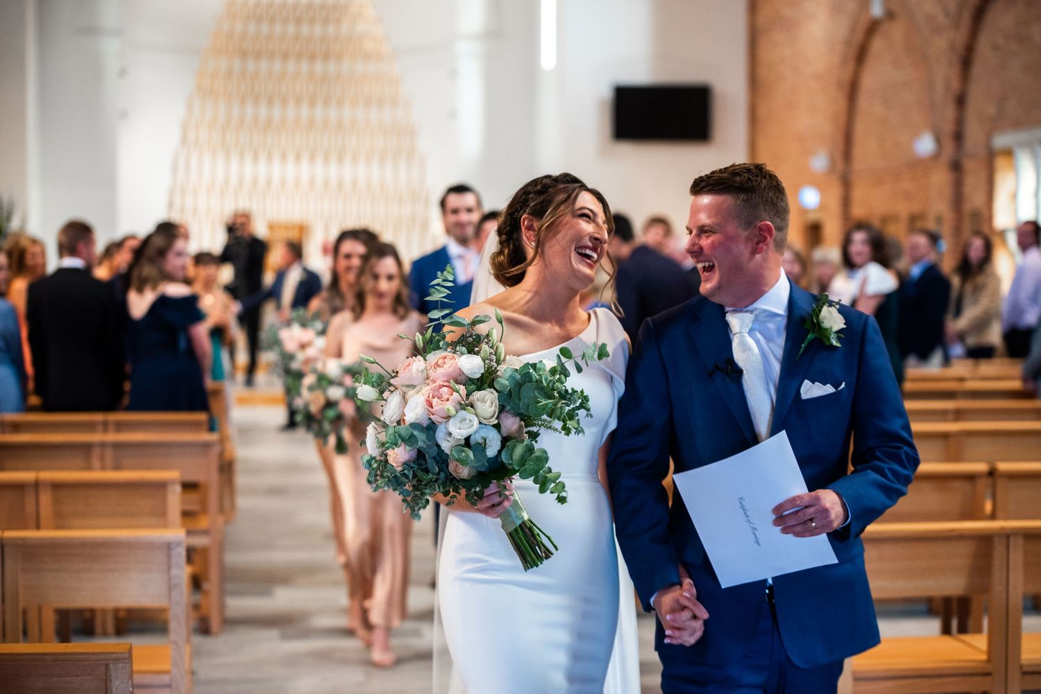 Wedding party walks down church aisle with bride and groom sharing a happy moment.