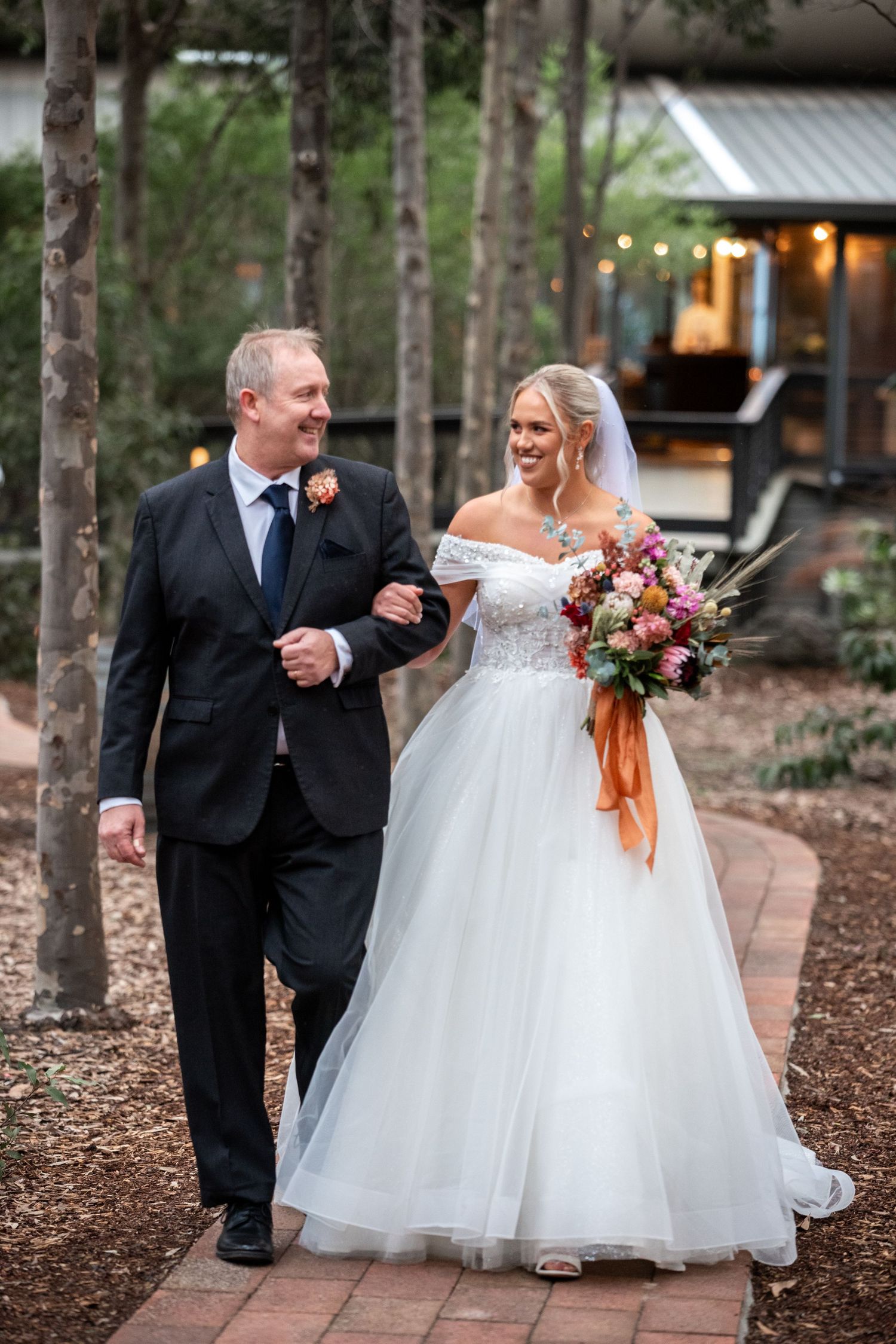 A bride in a white ball gown walks along a garden path during her outdoor wedding ceremony.
