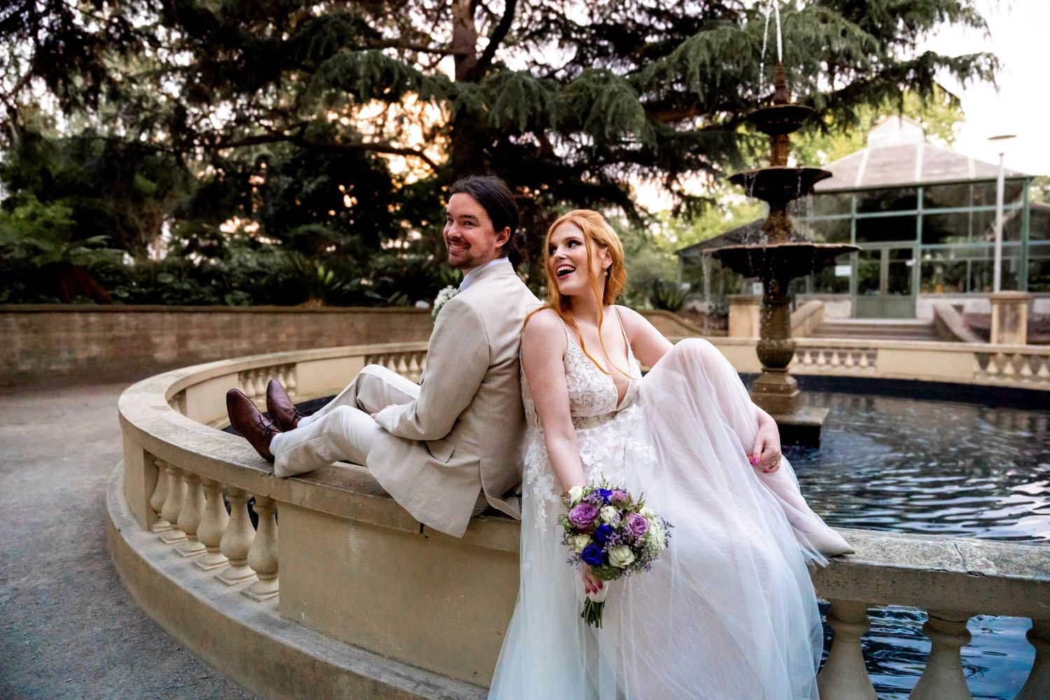Wedding couple sitting by a decorative stone fountain in a garden setting.