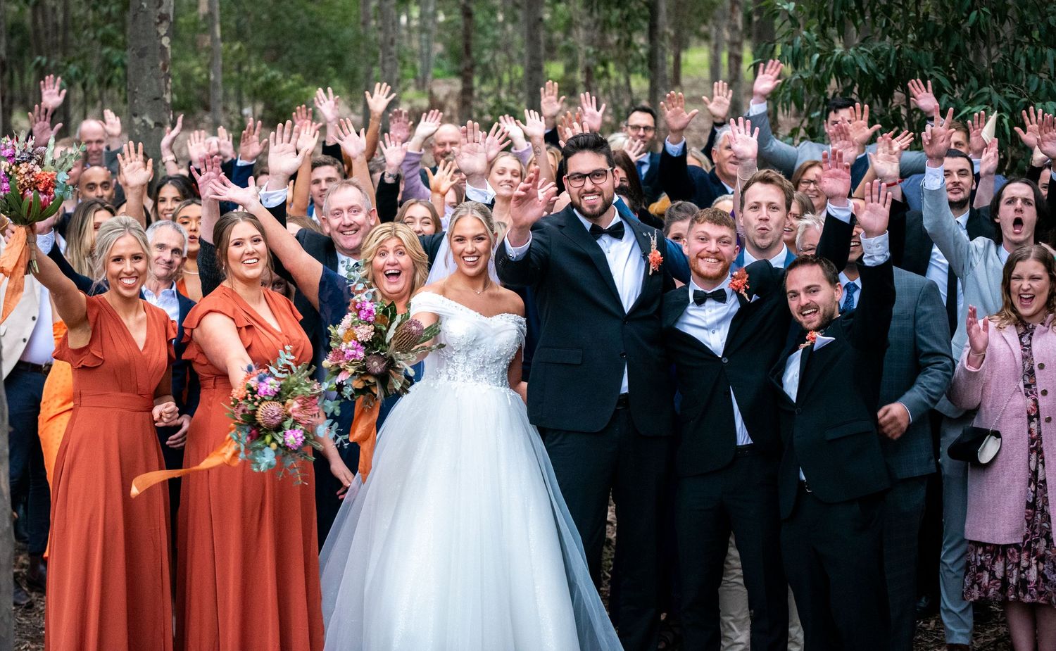 Large wedding party and guests pose for celebratory group photo outdoors among trees.