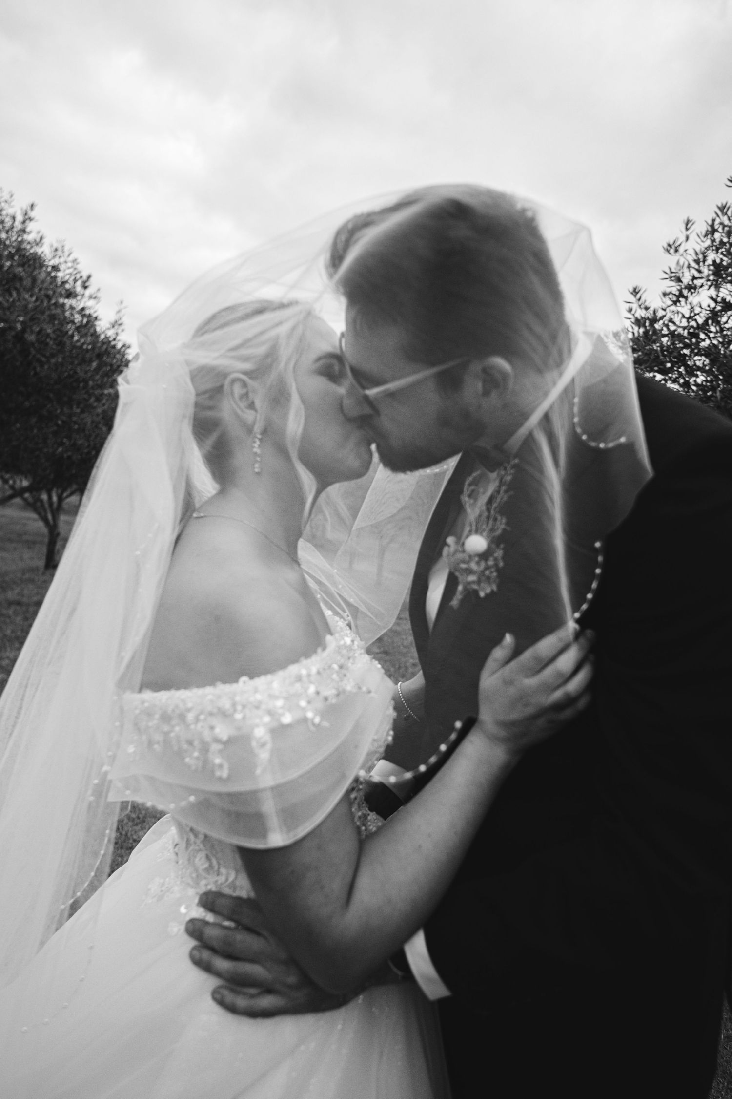 A romantic black and white wedding photo showing a tender kiss between newlyweds outdoors with a flowing veil.