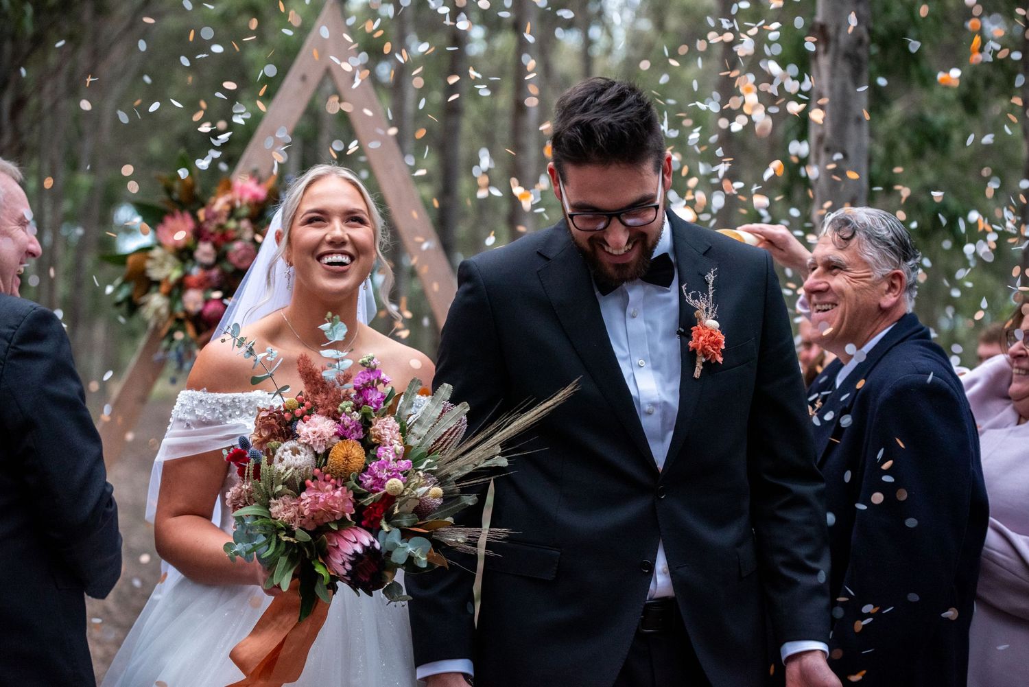 A joyful wedding couple exits their ceremony under falling confetti in an outdoor woodland setting.
