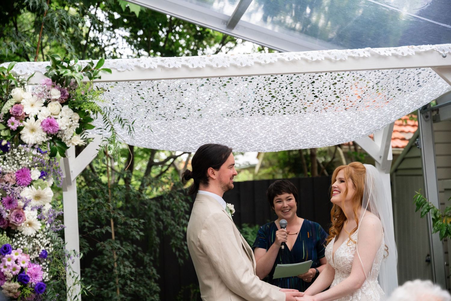 A couple shares a romantic moment under a floral-decorated white pergola during an outdoor wedding celebration.