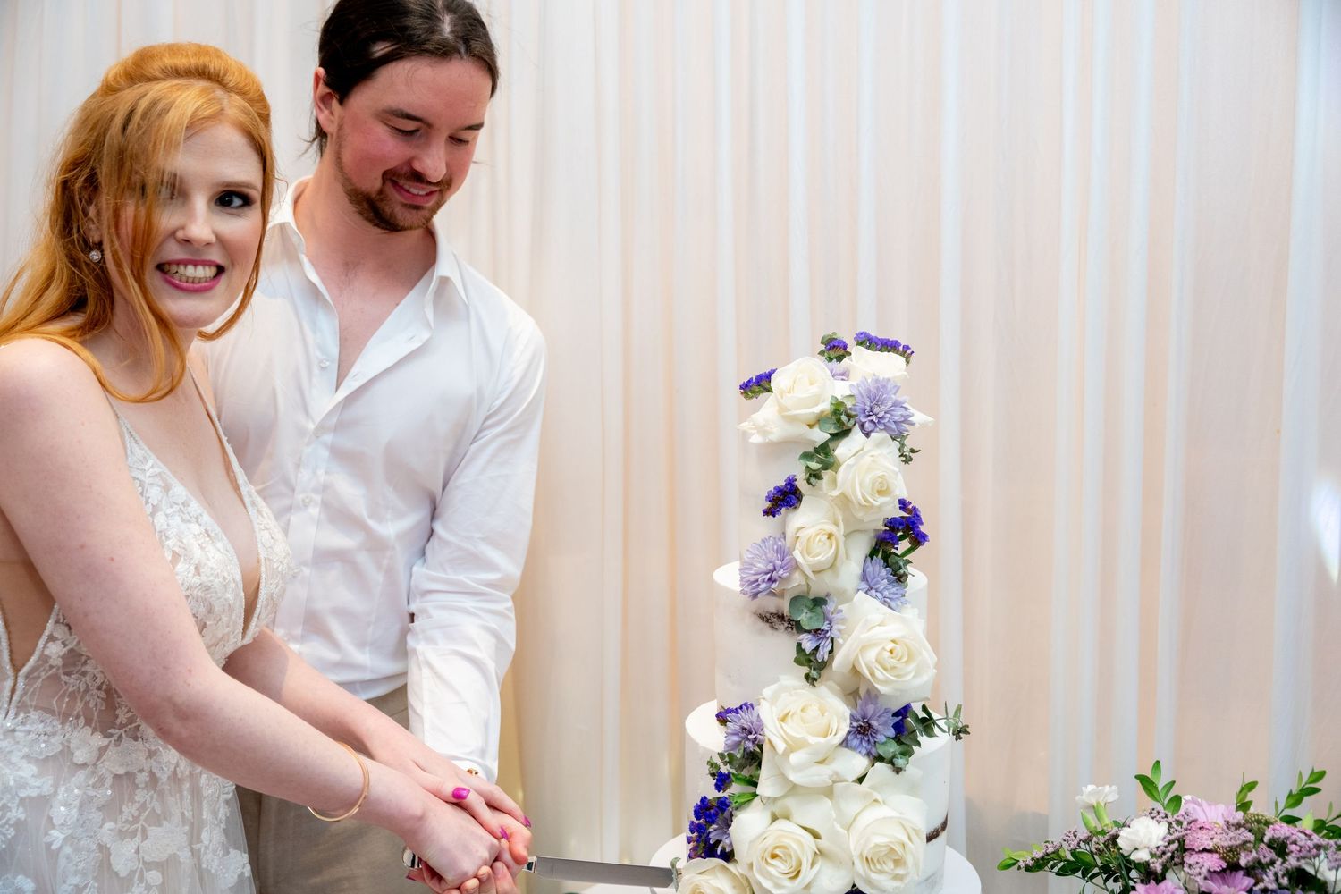 Couple cutting their elegant white wedding cake decorated with purple and blue flowers at reception.