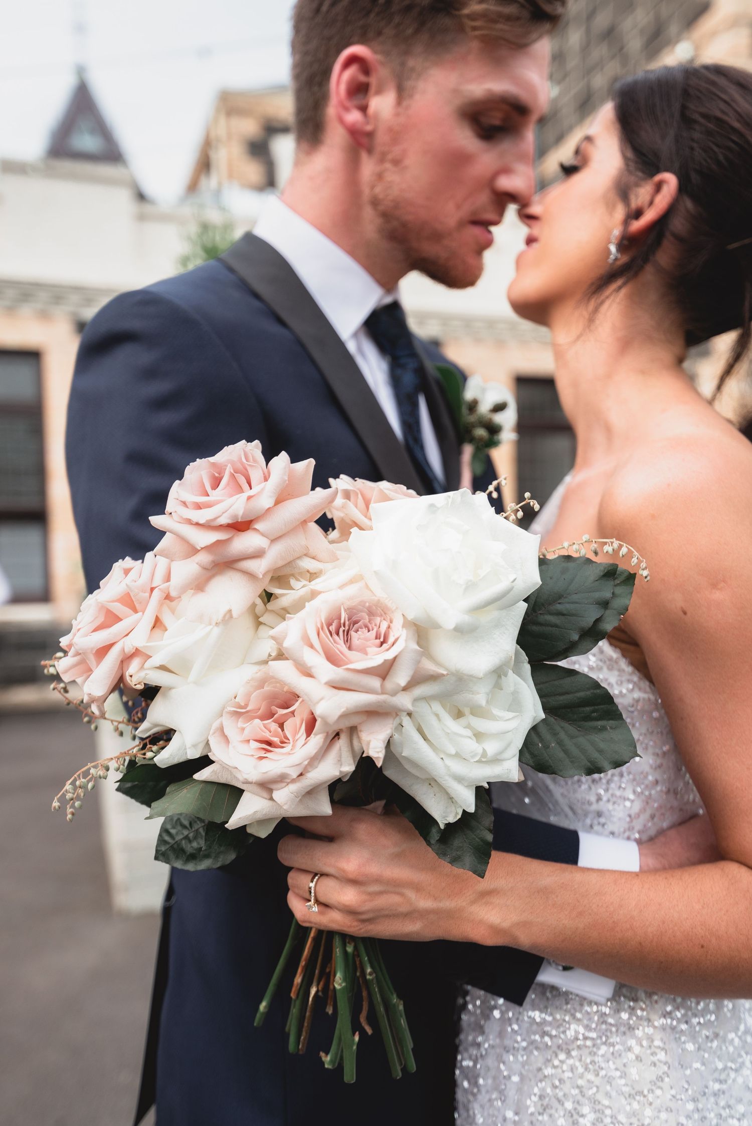 A romantic wedding couple embraces while holding a bouquet of pink and white roses against a blurred background.