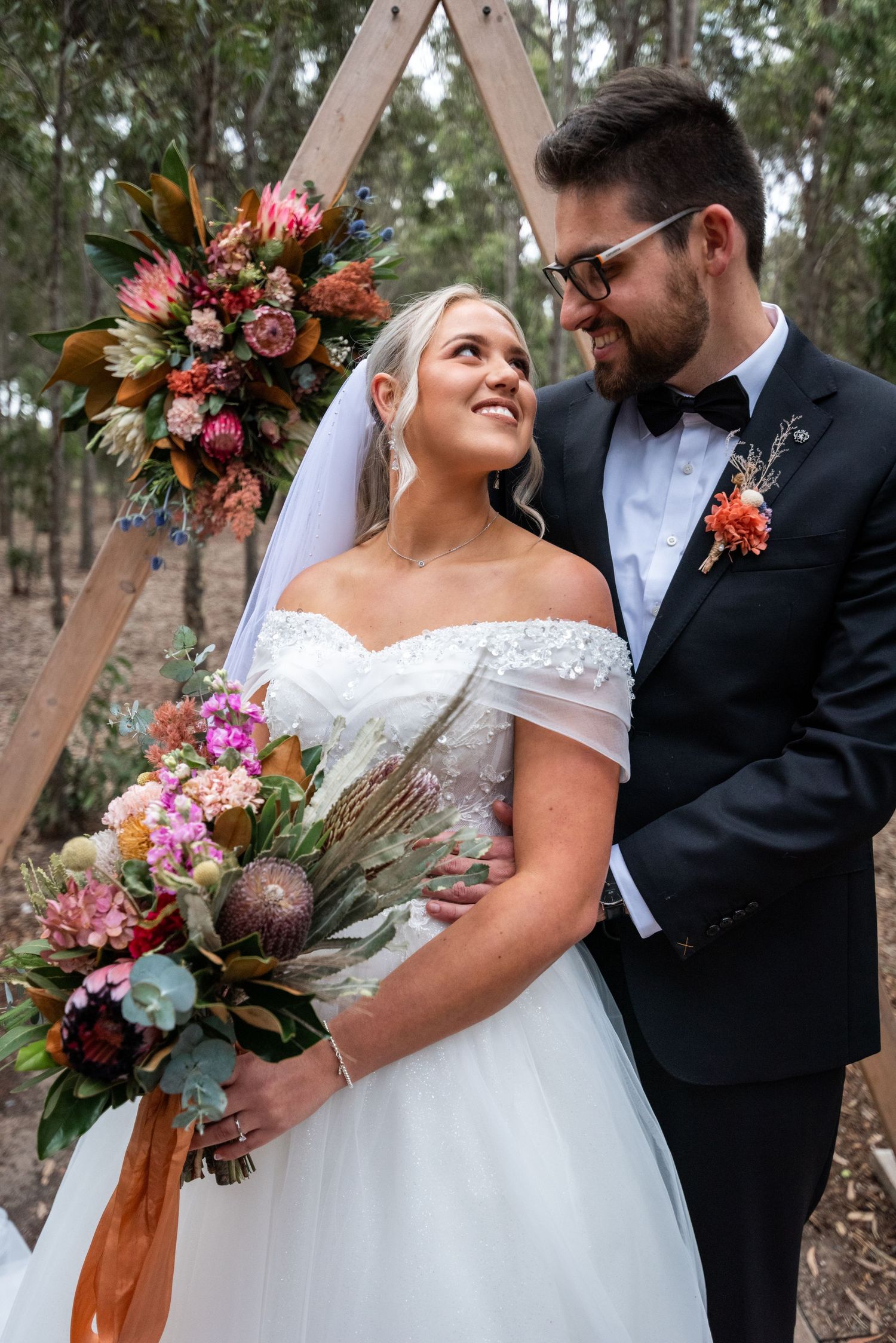 Wedding couple poses outdoors with vibrant autumn-colored bouquet against rustic wooden backdrop.