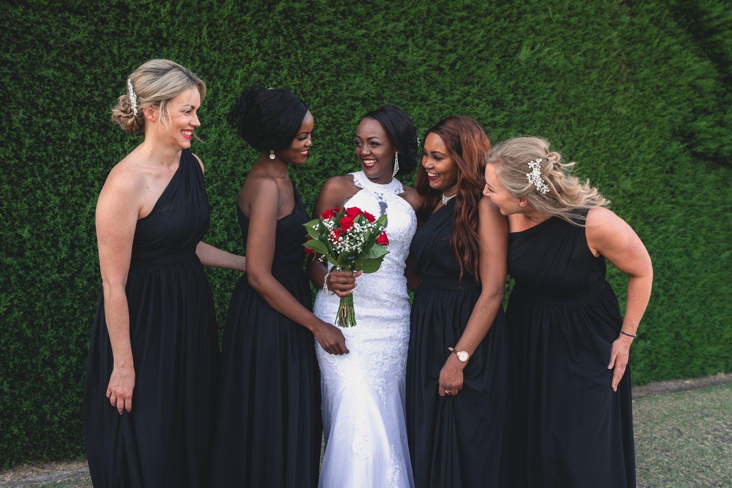 A wedding party wearing black and white dresses stands in a line outdoors against a green hedge background.