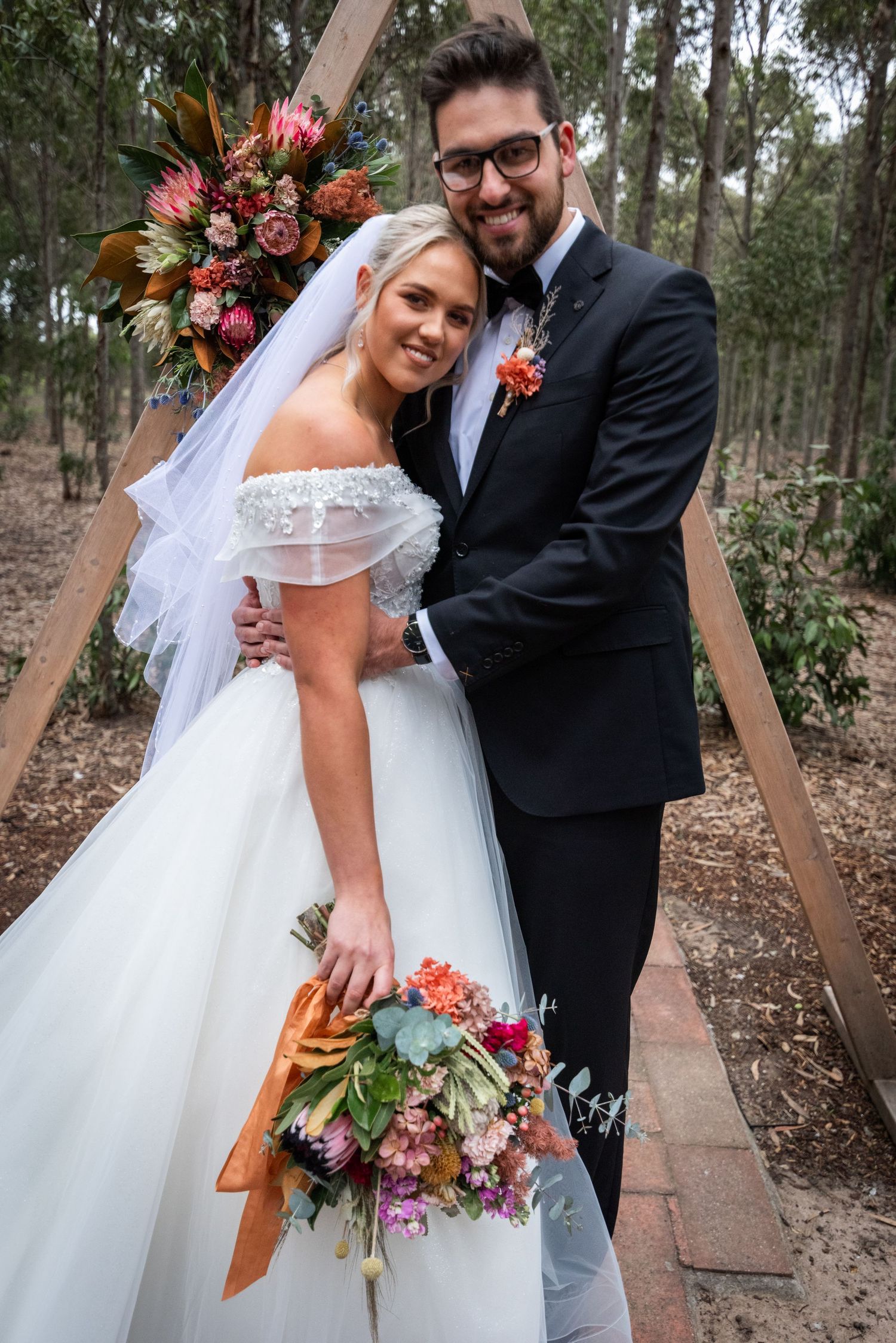A newlywed couple poses on a wooden walkway surrounded by trees, with the bride holding a vibrant autumn bouquet.