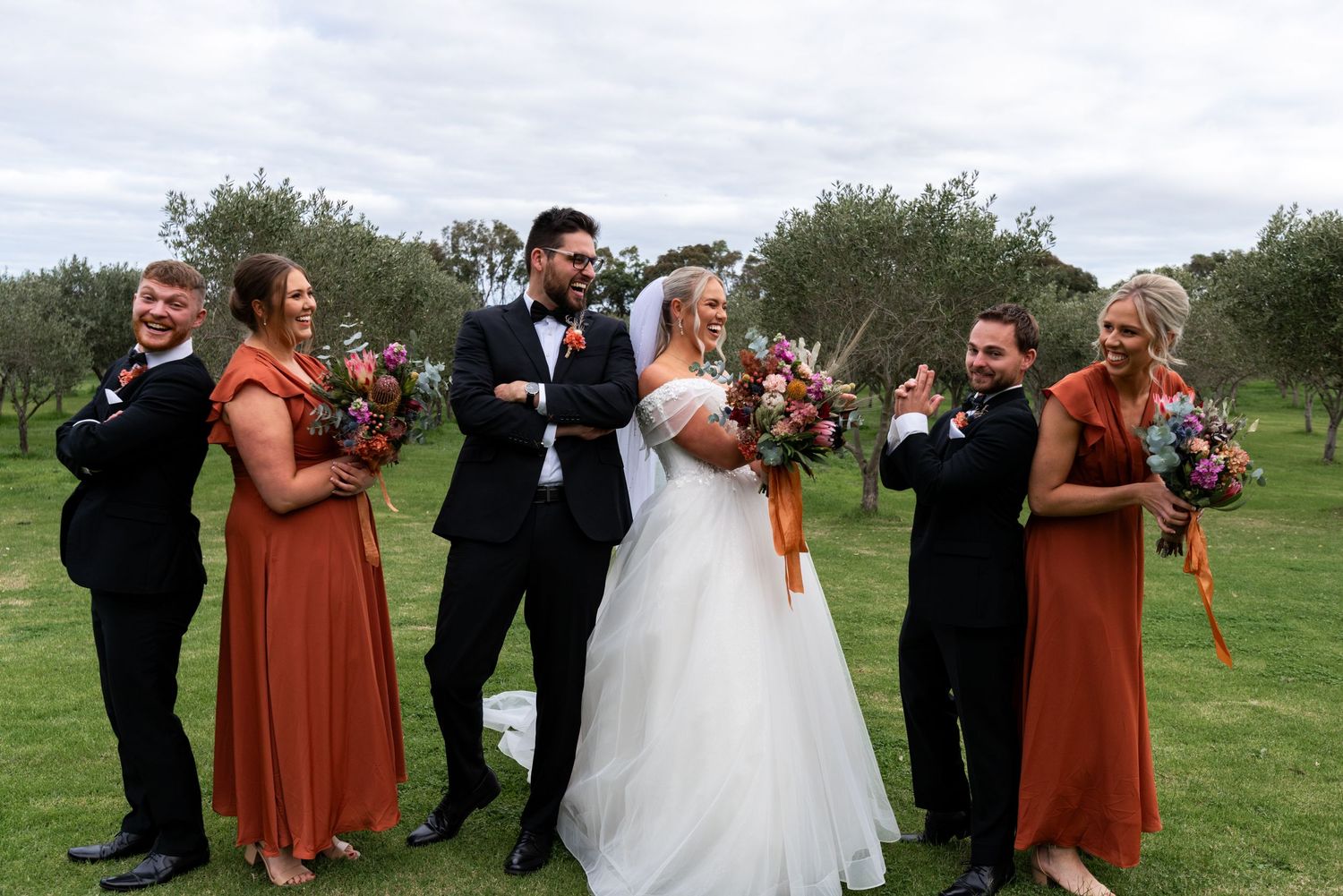 Wedding party in black suits and rust dresses pose together on grass.