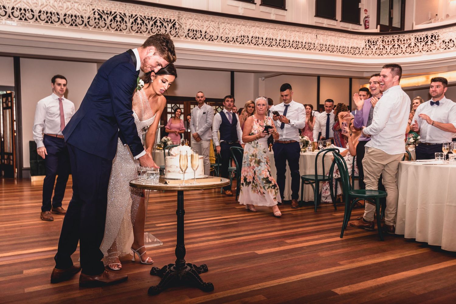 A couple shares a romantic moment while cutting their wedding cake in an elegant reception hall with wooden floors and guests watching.