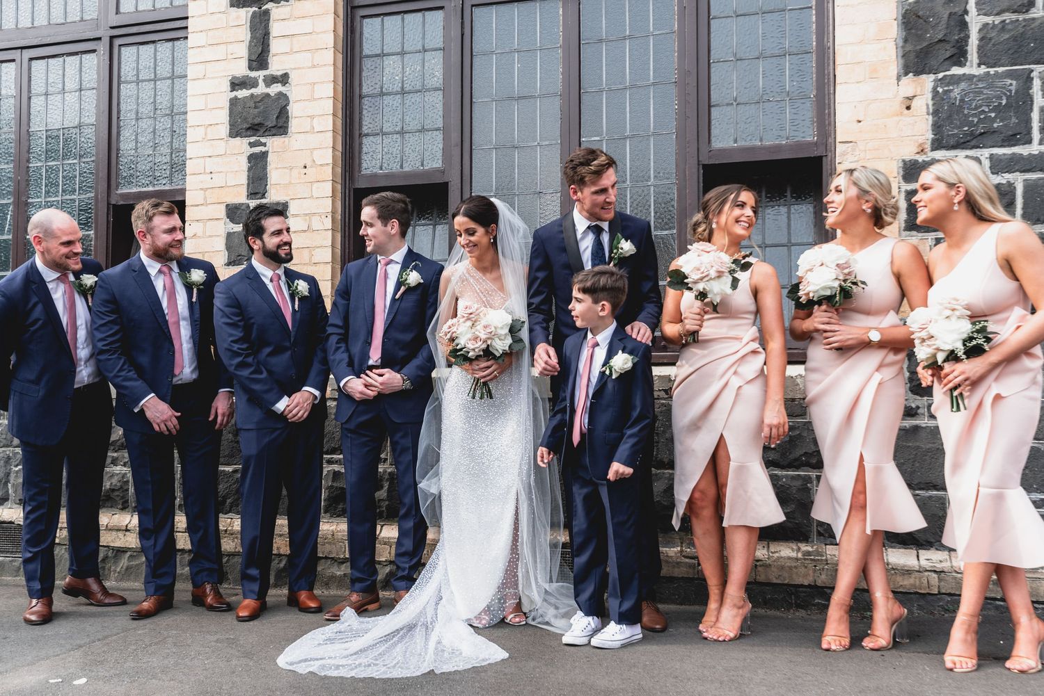 Wedding party in formal attire poses outside stone building with bridesmaids in blush pink dresses and groomsmen in navy suits.