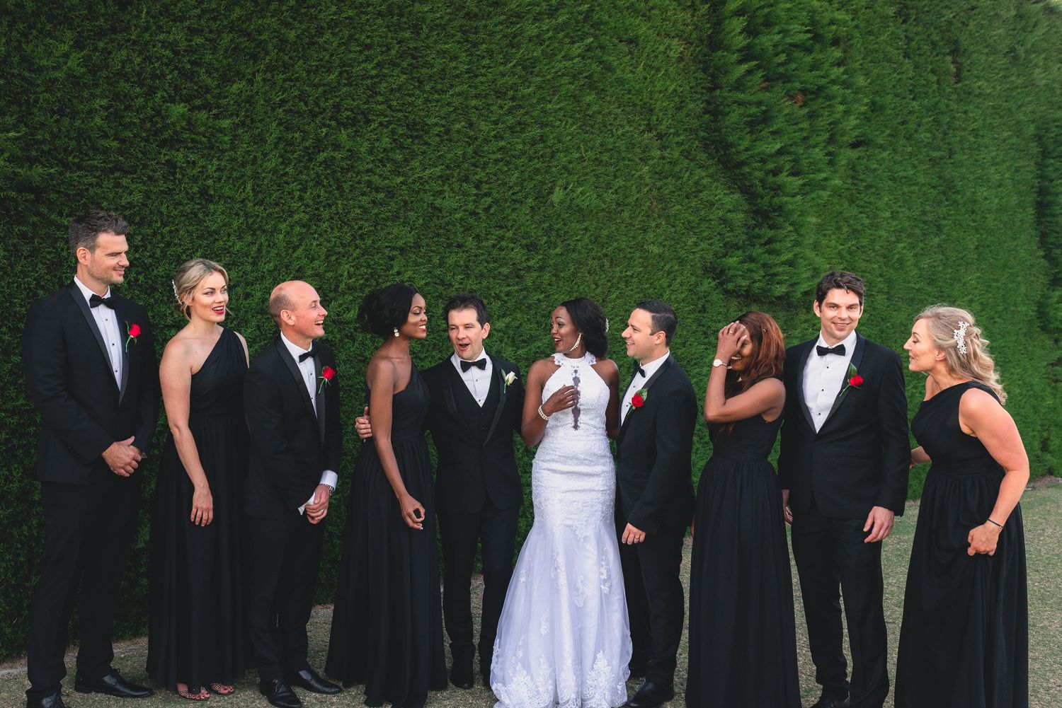 Wedding party in formal attire laughs together while standing against a lush green hedge wall backdrop.