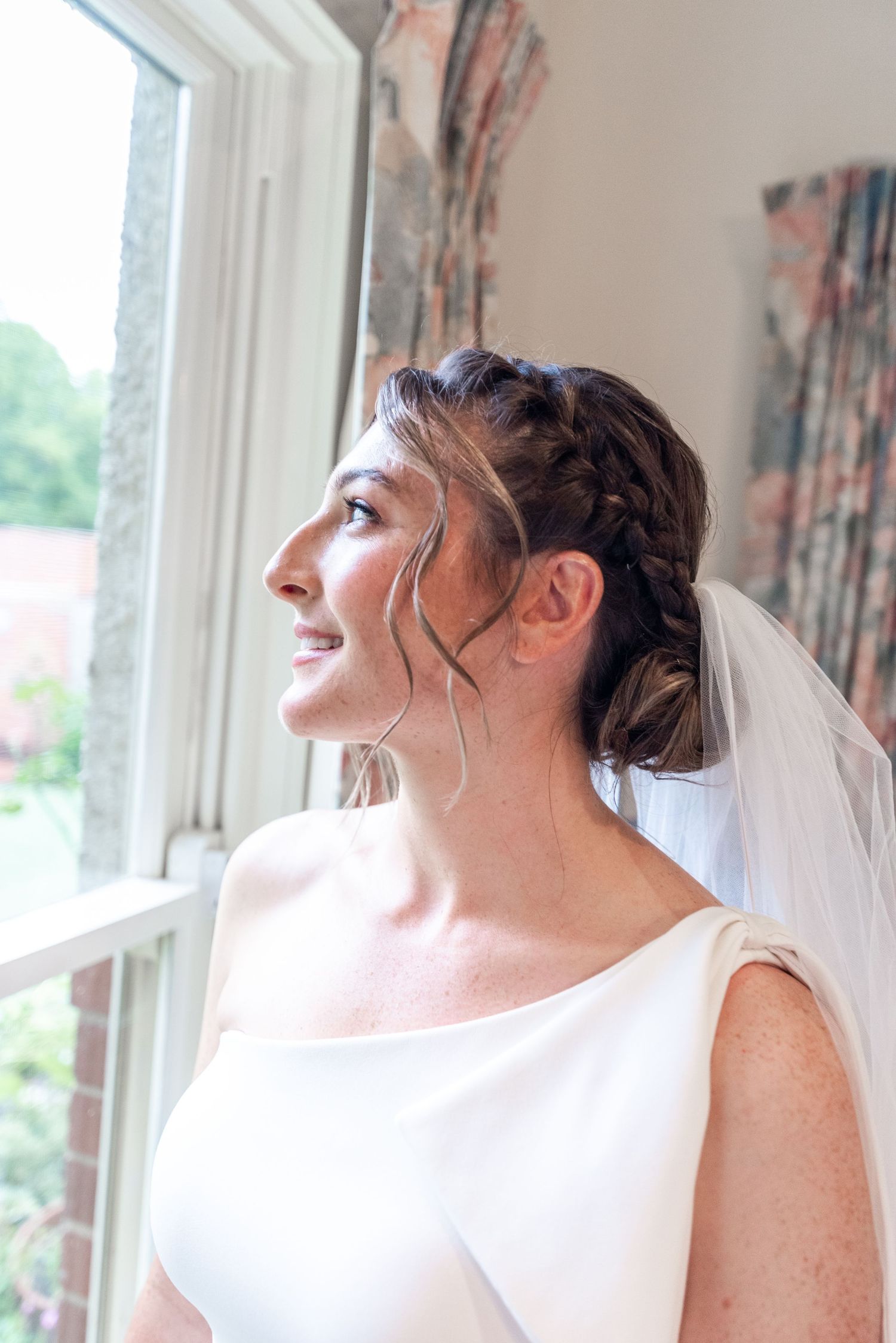 Bride in white dress laughs joyfully while looking upward near windows with floral curtains.