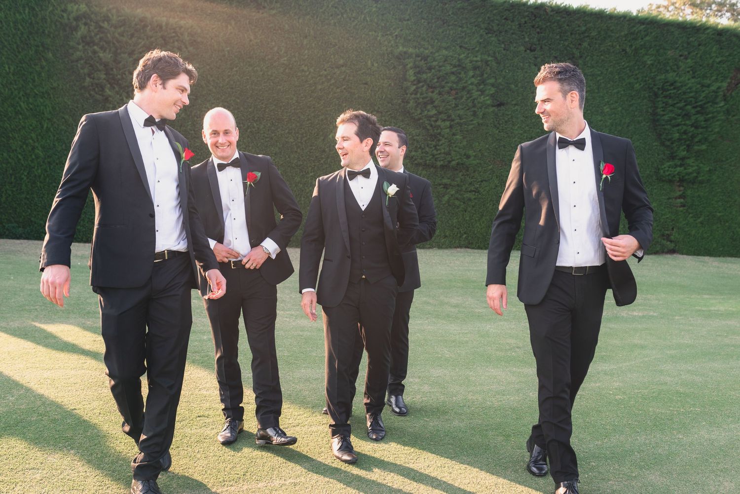 A sequence of groomsmen in black tuxedos walking in sync on a green lawn during a wedding photoshoot.