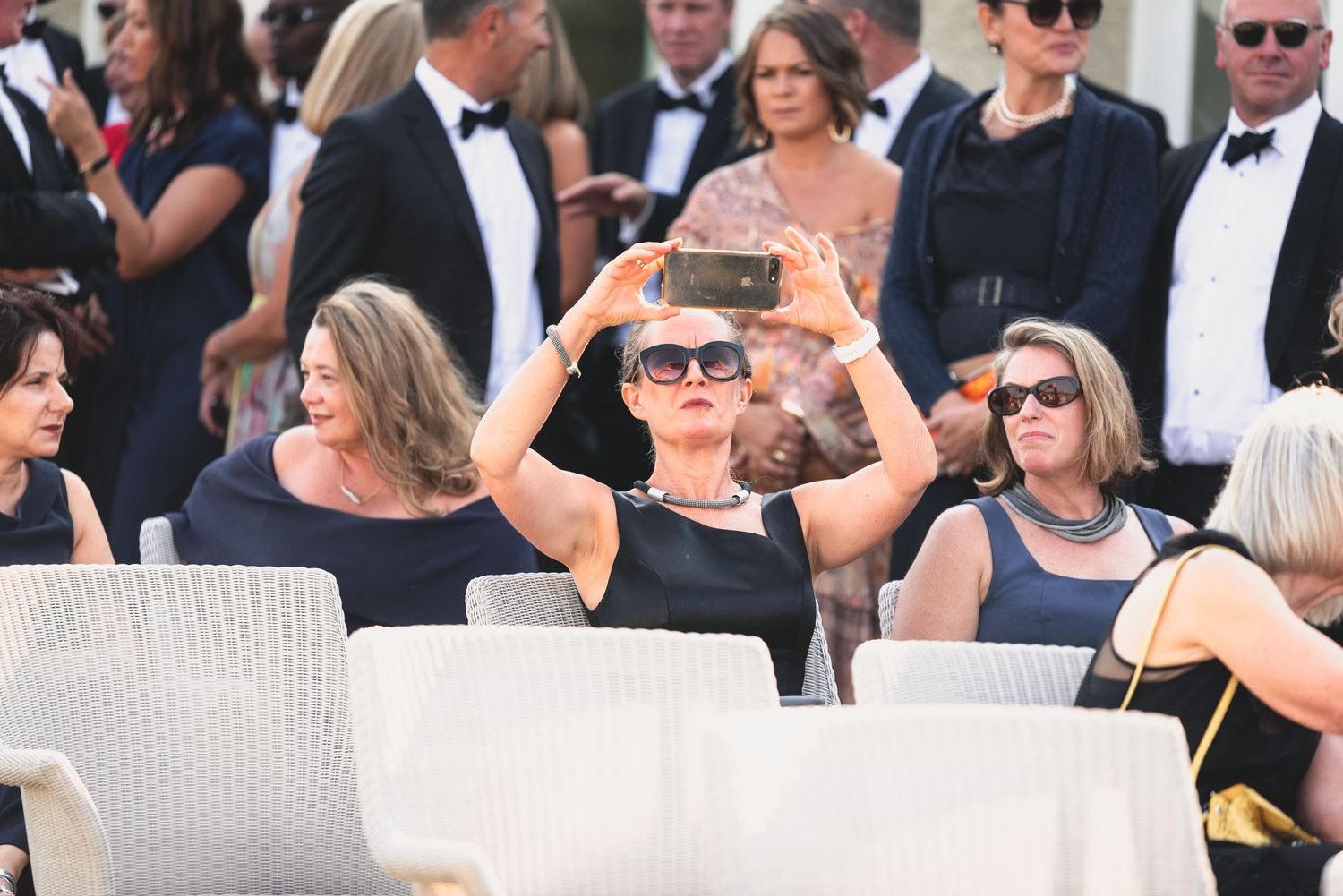 Crowd of formally dressed people attending an outdoor event with some wearing sunglasses and dark attire.