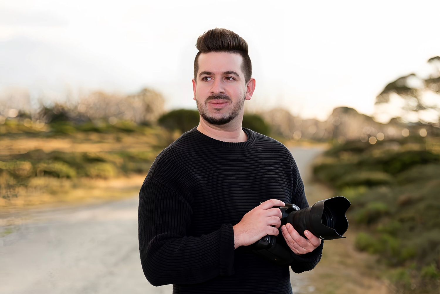 A photographer wearing a black sweater holds a camera while standing on a rural road at sunset.