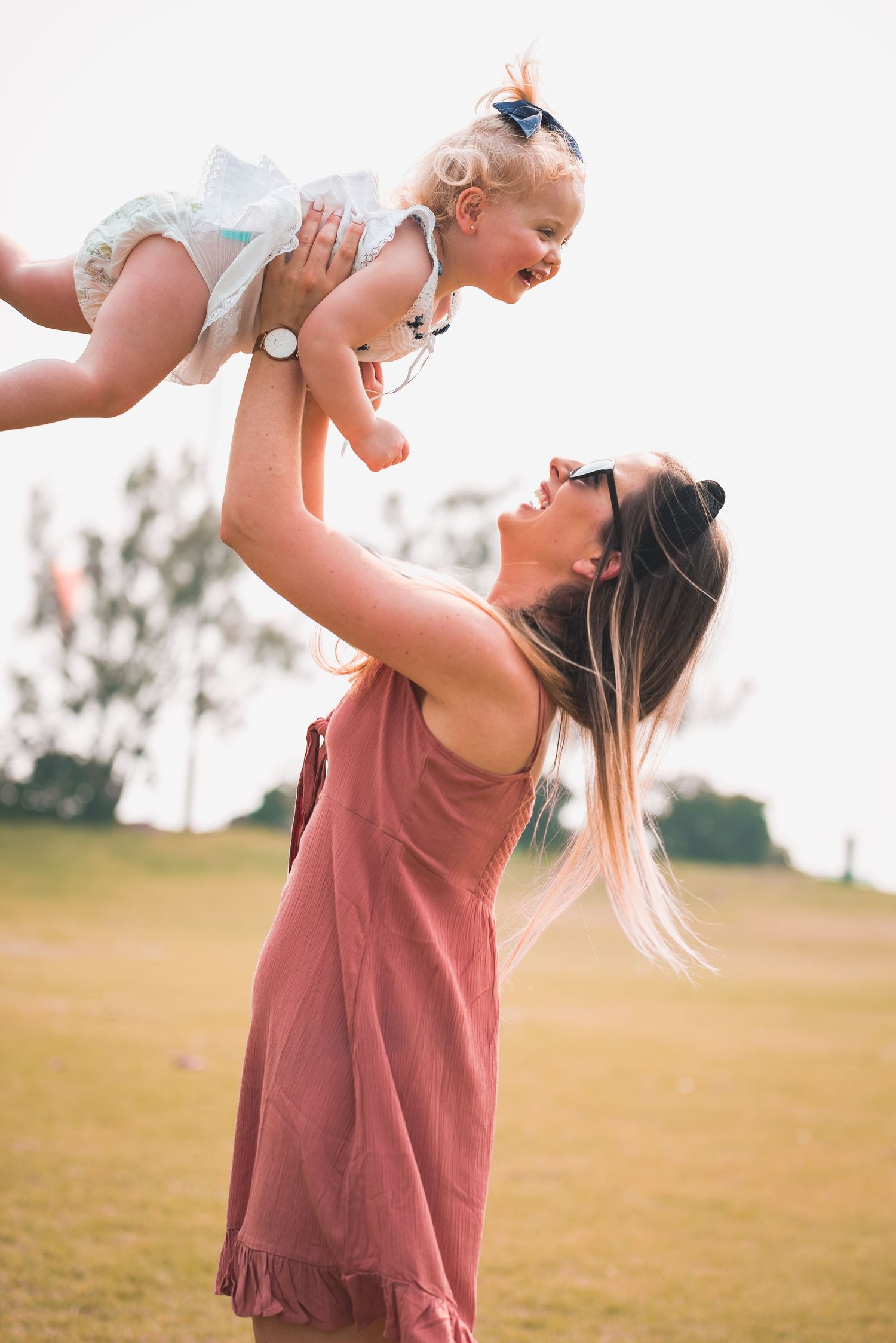 A person in a mauve dress lifts and plays with a baby outdoors in a grassy field during golden hour.