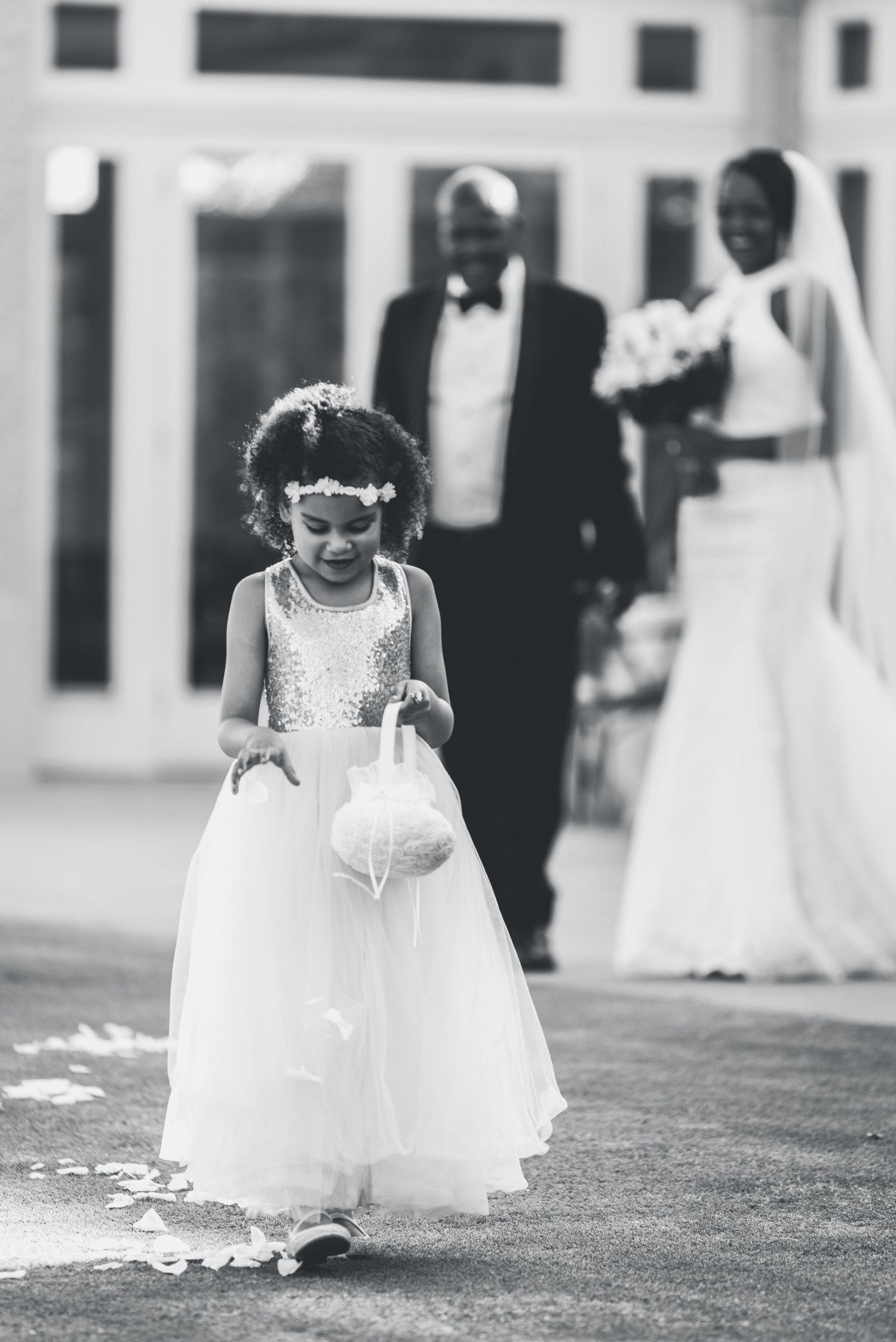 A flower girl in a white tulle dress carries a basket while walking down the aisle at a formal wedding ceremony.