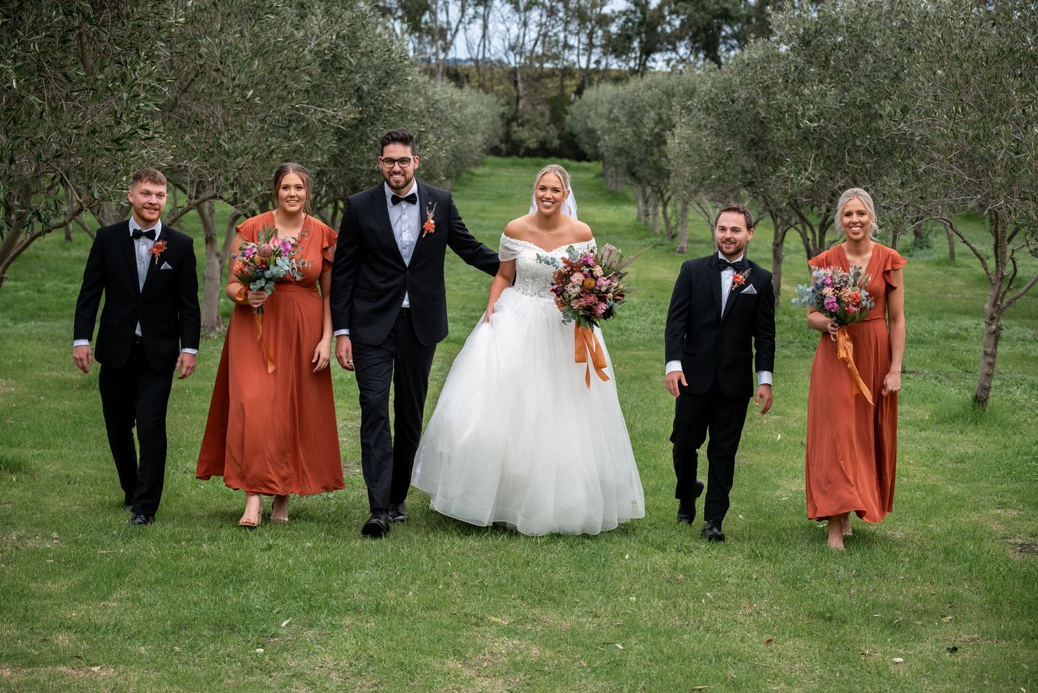 Wedding party walks together on grassy field with bridesmaids in burnt orange dresses.