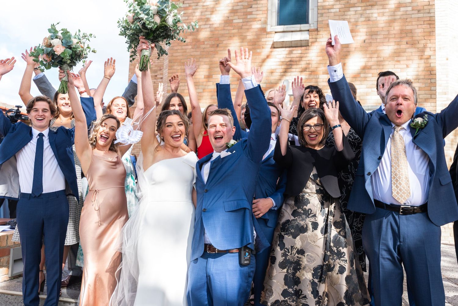 Wedding party celebrates joyfully with raised arms outside brick church on sunny day.