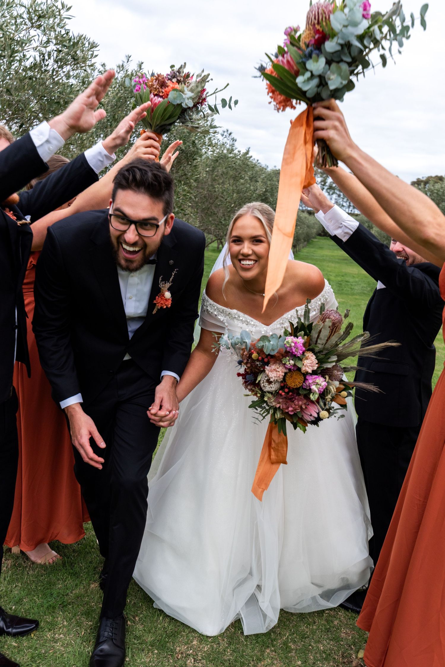 Wedding party celebrates outdoors with joyful dancing and colorful burnt orange bridesmaid dresses.