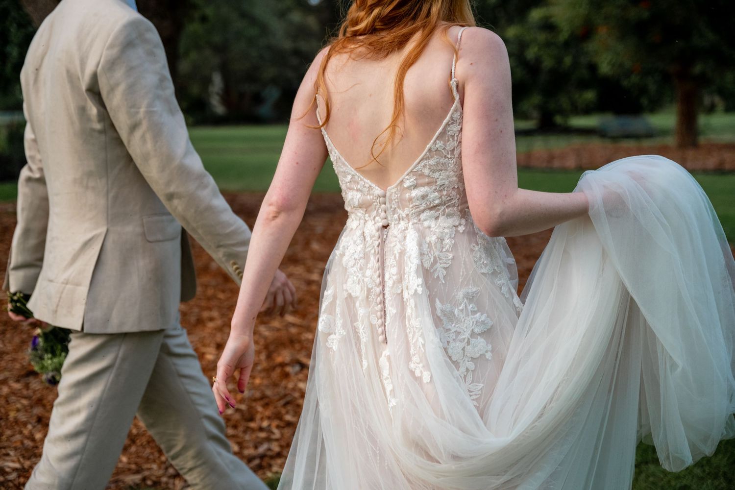 A bride in a lace wedding dress walks through autumn leaves with someone in a light beige suit.