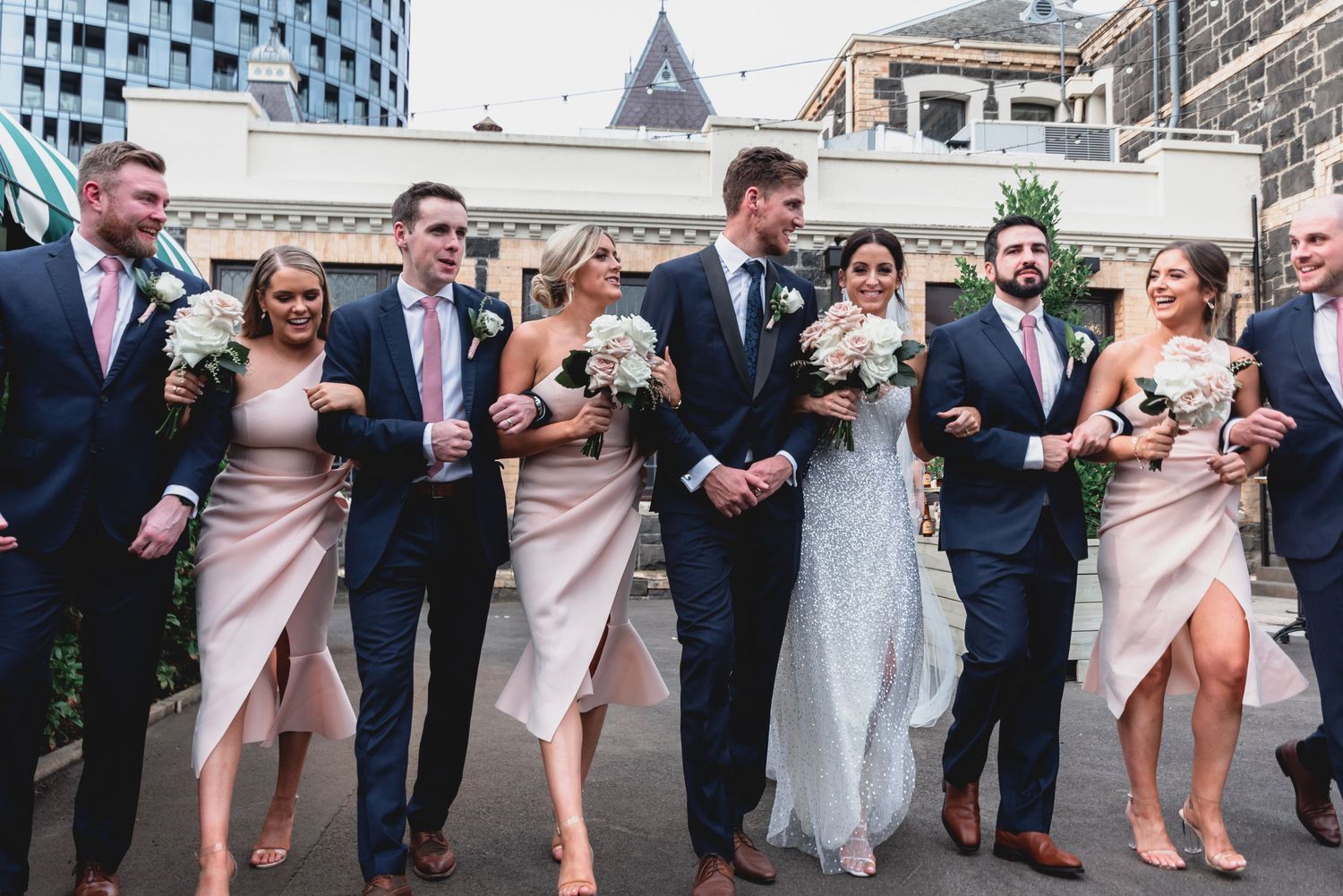 Wedding party in blush pink dresses and navy suits walk and laugh together outdoors.
